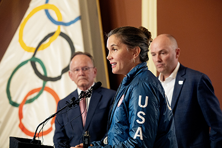 Salt Lake City Mayor Erin Mendenhall answers questions at a microphone in front of the Olympic flag in the atrium of City Hall. Behind her stand Utah Senate President Stuart Adams, left, and Utah Governor Spencer Cox, right.