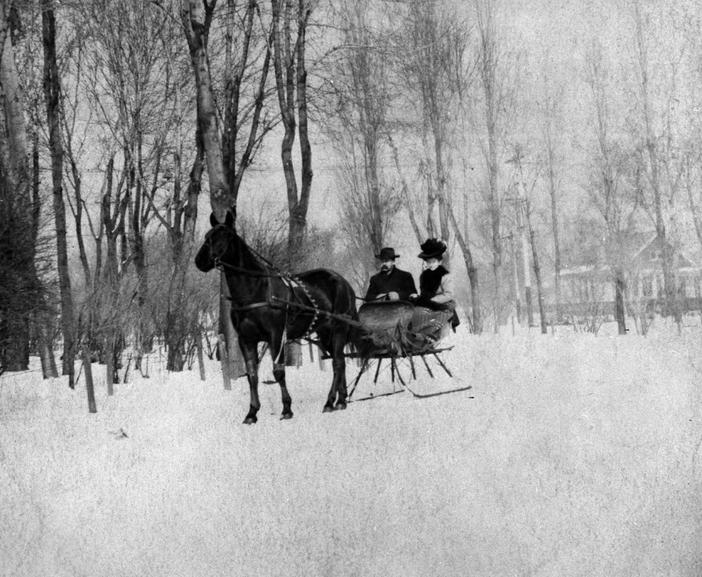Historical black and white photo of a man and a woman riding in a sleigh pulled by a black horse, with liberty park covered in snow