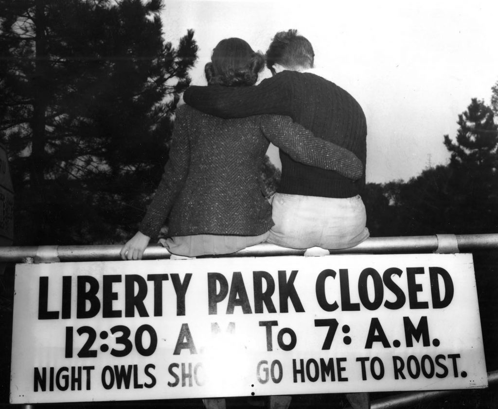 Historical black and white photo of two people with their arms around each other, sitting on a railing above a sign. The sign reads "Liberty Park Closed 12:30 AM to 7 AM. Night owls should go home to roost." in all capitals