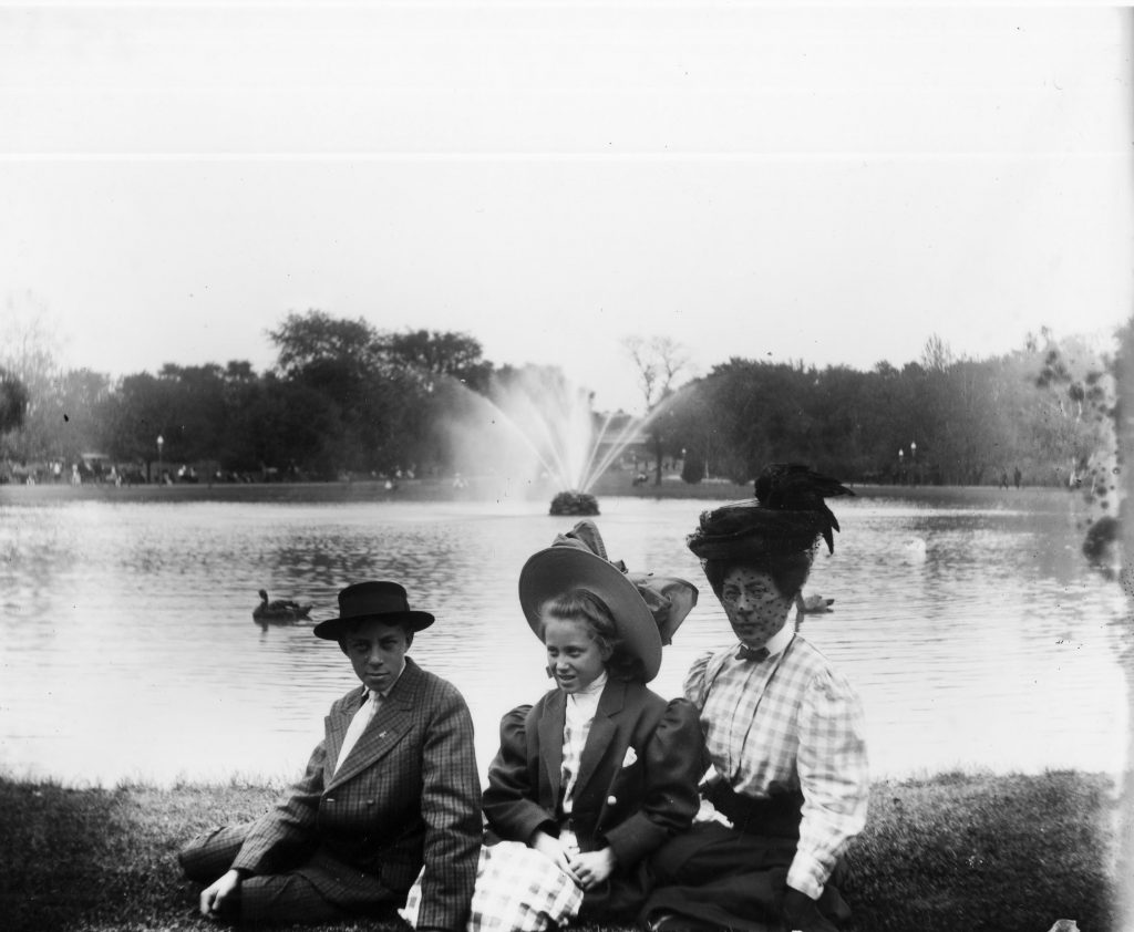 Historical black and white photo showing three people sitting on the grass by the liberty park pond, likely taken in the late 1800s or early 1900s 
