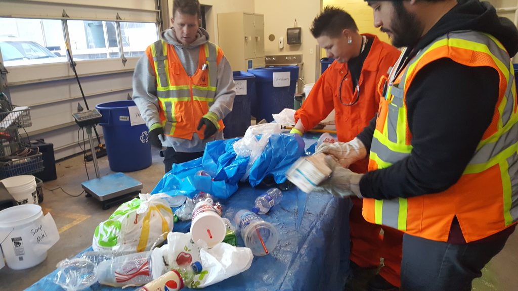 Three Waste and Recycling Department workers sort through trash on a table.