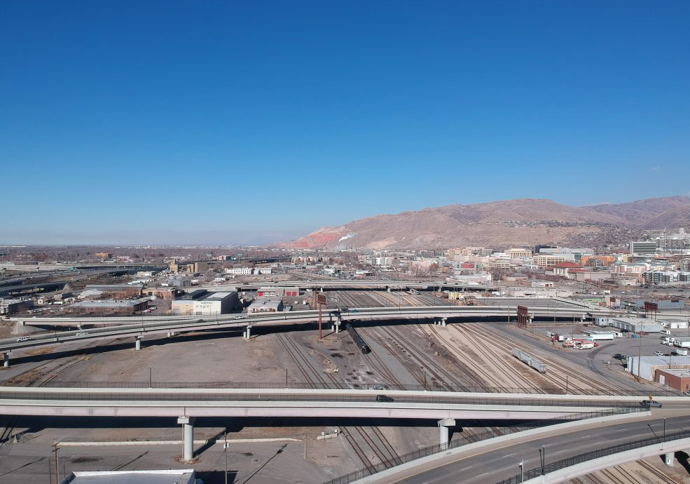 An aerial view of Salt Lake City showing the east-west transportation divide created by the railroad and I-15 freeway. Multiple elevated highways and rail lines cut through the city, separating neighborhoods and limiting connectivity.
