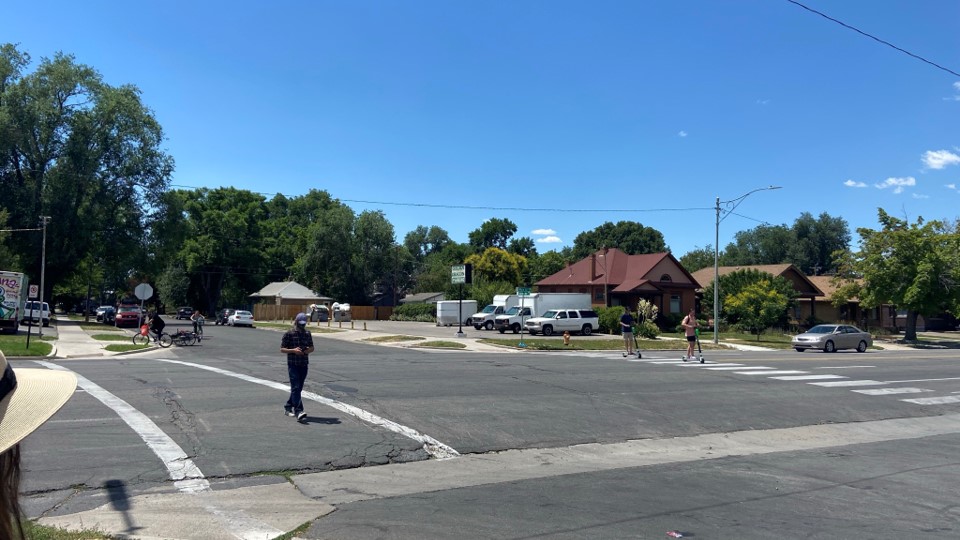 People walking and biking across a wide, marked crosswalk on Main Street in Salt Lake City, with houses, trees, and parked cars in the background on a sunny day.