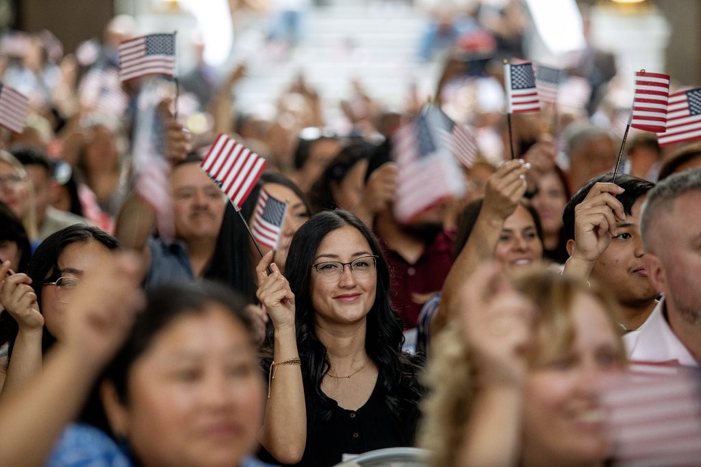 A smiling woman with glasses holding an American flag among a crowd waving flags.
