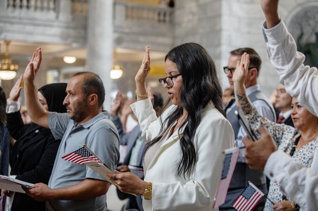 A group of people, with one woman with glasses in focus, raising her hand while taking the Oath of Alliance at a citizenship ceremony.