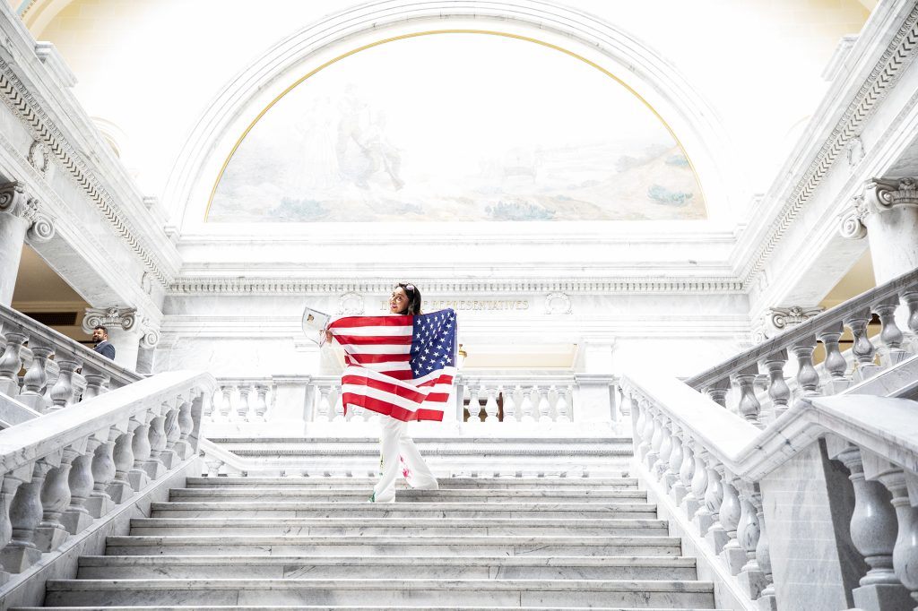 A woman holding a large U.S. flag behind her while standing on the stairs inside the Utah Capitol building.