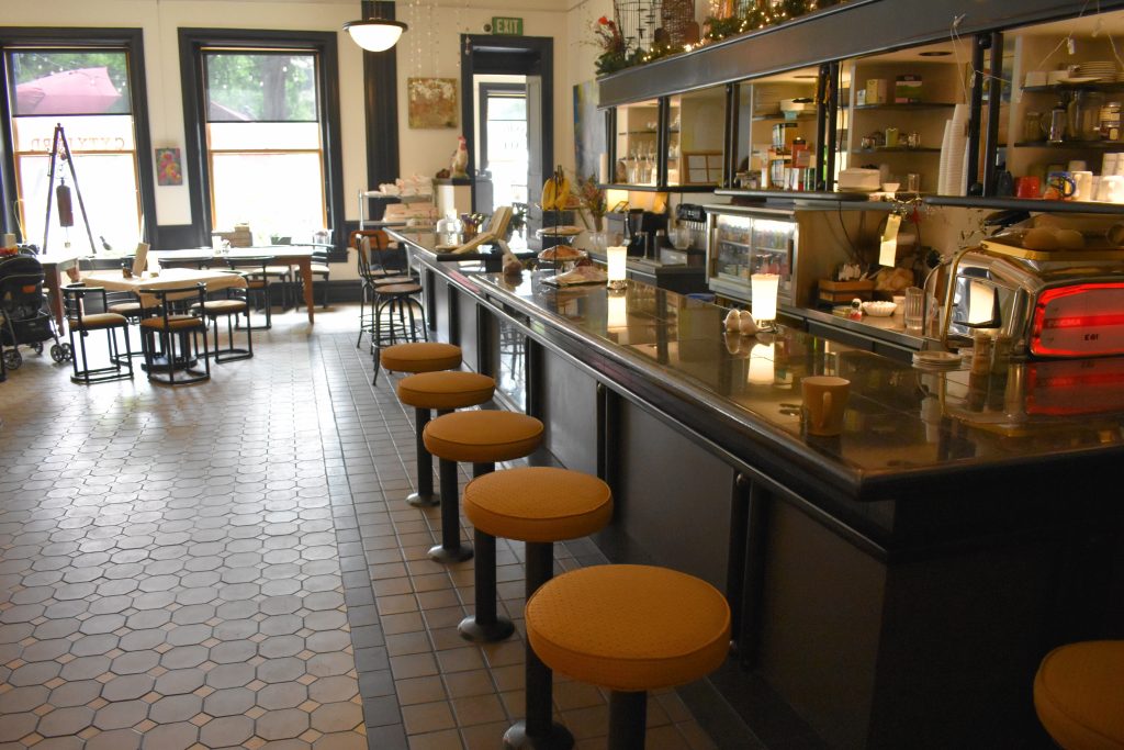 Photo of the interior of the cafe. Where are stools lined up at a bar, and tables and chairs in the room that is dimly lit with large windows on the far wall.