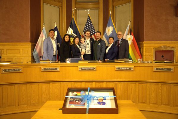 A group of eight people in formal attire stand behind a desk with flags in the background, in the council chamber. Council Member Chris Wharton is being recognized for his leadership as the council chair in 2025.