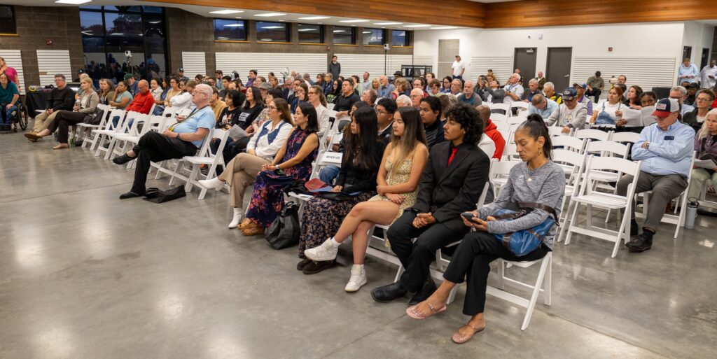 A large audience sits in rows of white folding chairs in a brightly lit indoor meeting room.