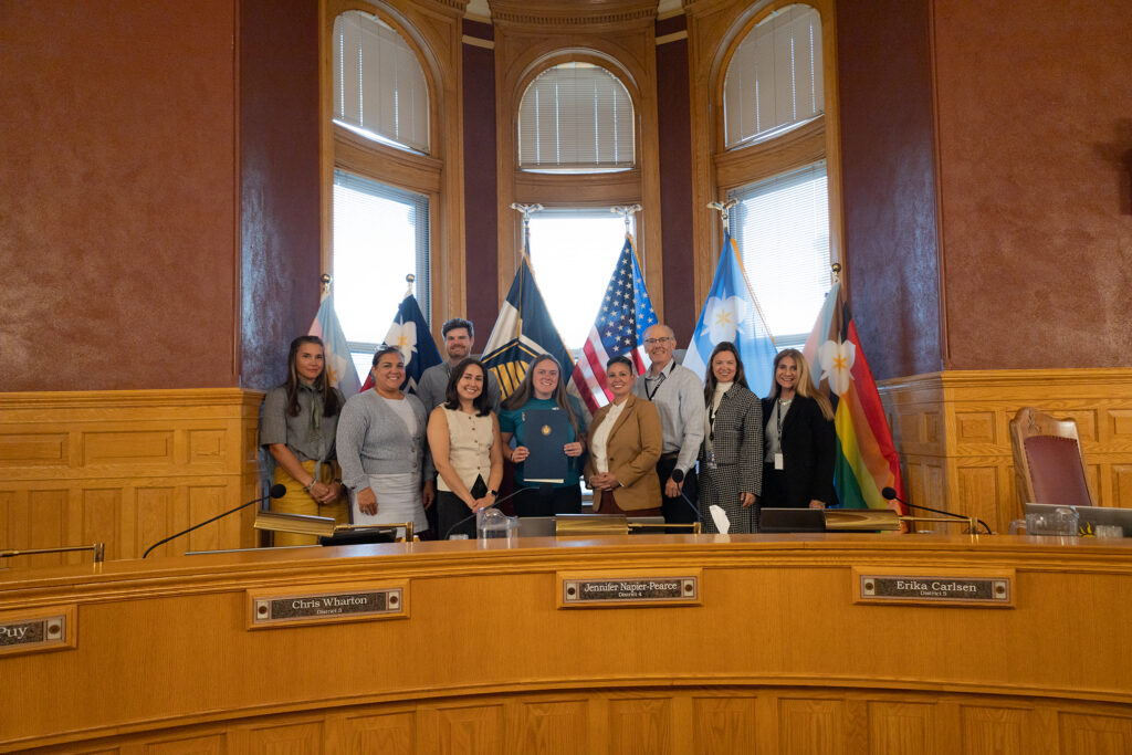 Eight people pose in a city council chamber with flags as one person holds a ceremonial resolution recognizing National Suicide Prevention Month.