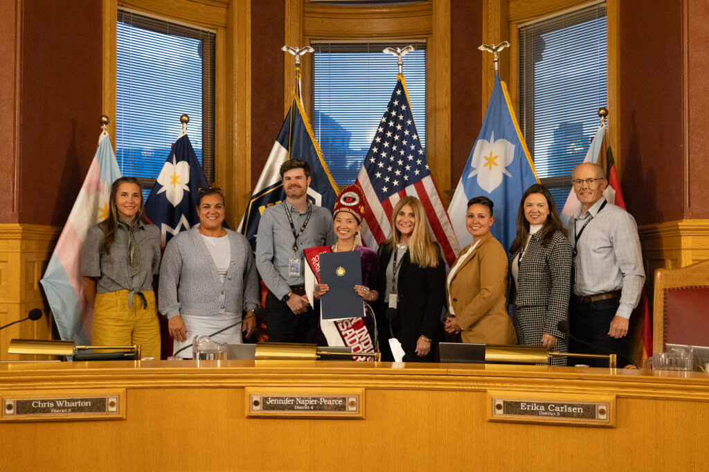 Eight people pose in a city council chamber with flags as one person holds a ceremonial resolution declaring Indigenous Peoples Day.