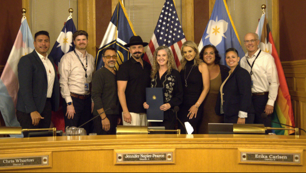 Nine people pose in a city council chamber with flags as one person holds a ceremonial resolution recognizing Italian American Heritage Month.