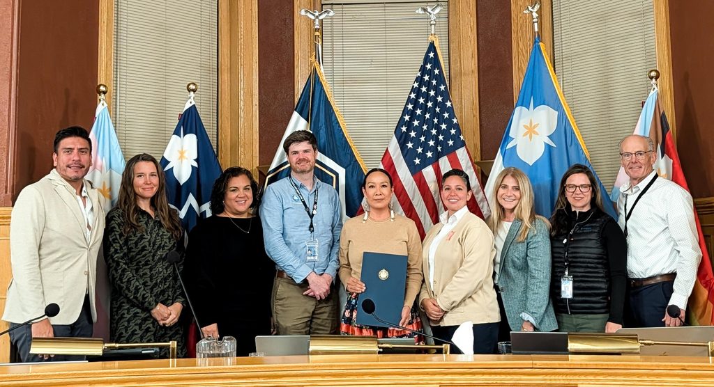 The Salt Lake City Mayor and City Council presenting a ceremonial resolution. Nine people standing in front of flags while someone holds the resolution.