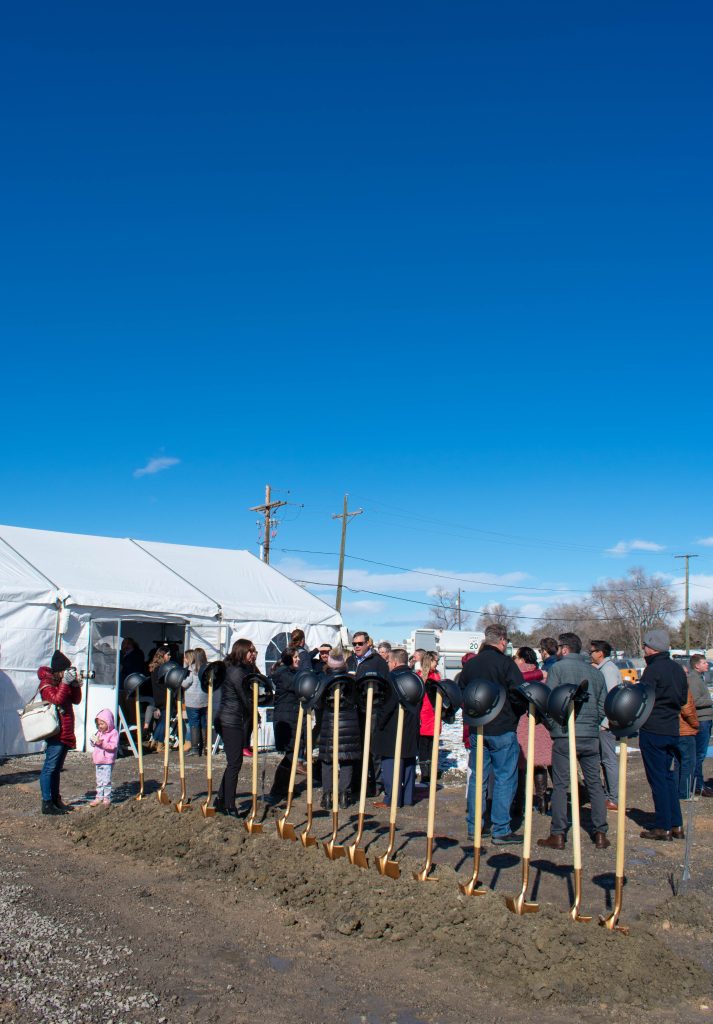 15 shovels stuck straight up in  the dirt with hard hats on top of the handles with a large group of people talking behind them.