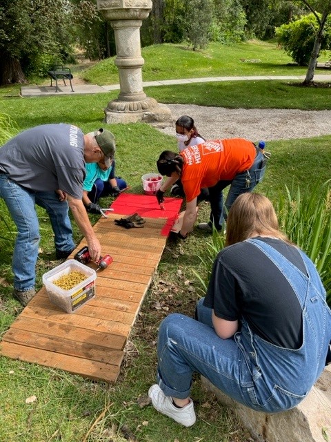 People painting a small bridge, that is over a tiny section of the man-made river, red.