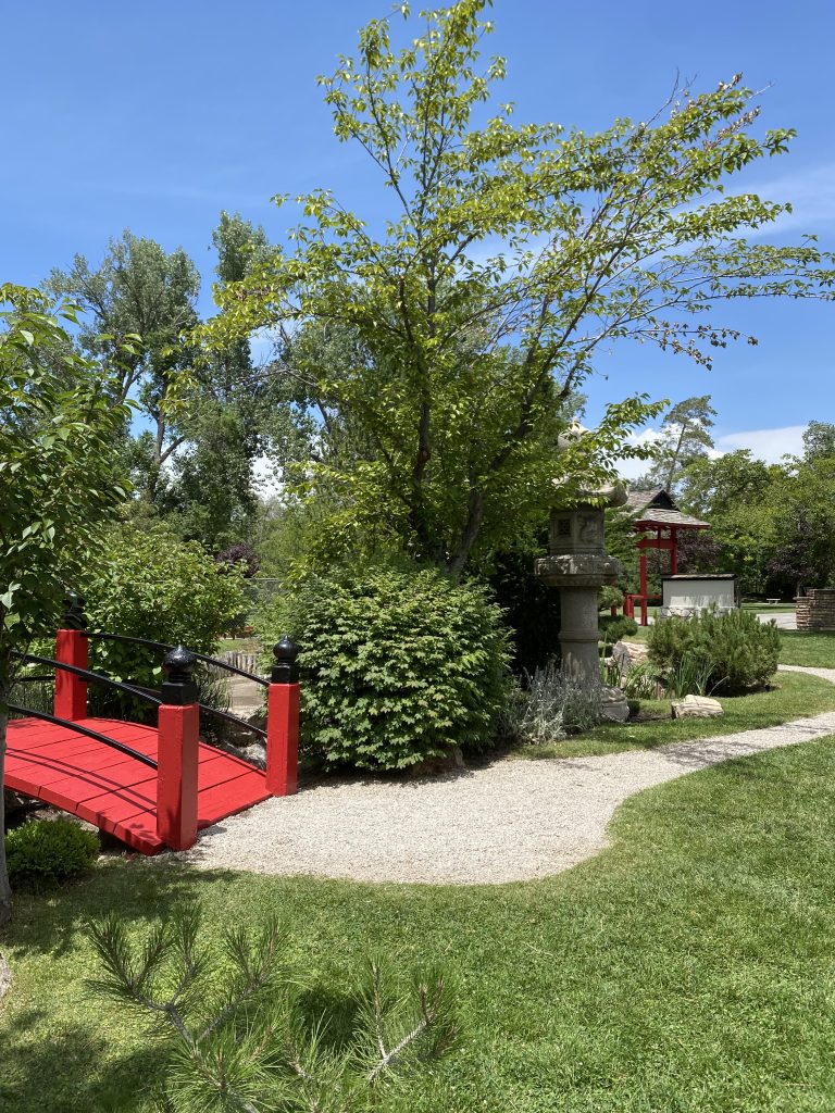 A red bridge with a light stone path leading to it and trees and bushes around it.