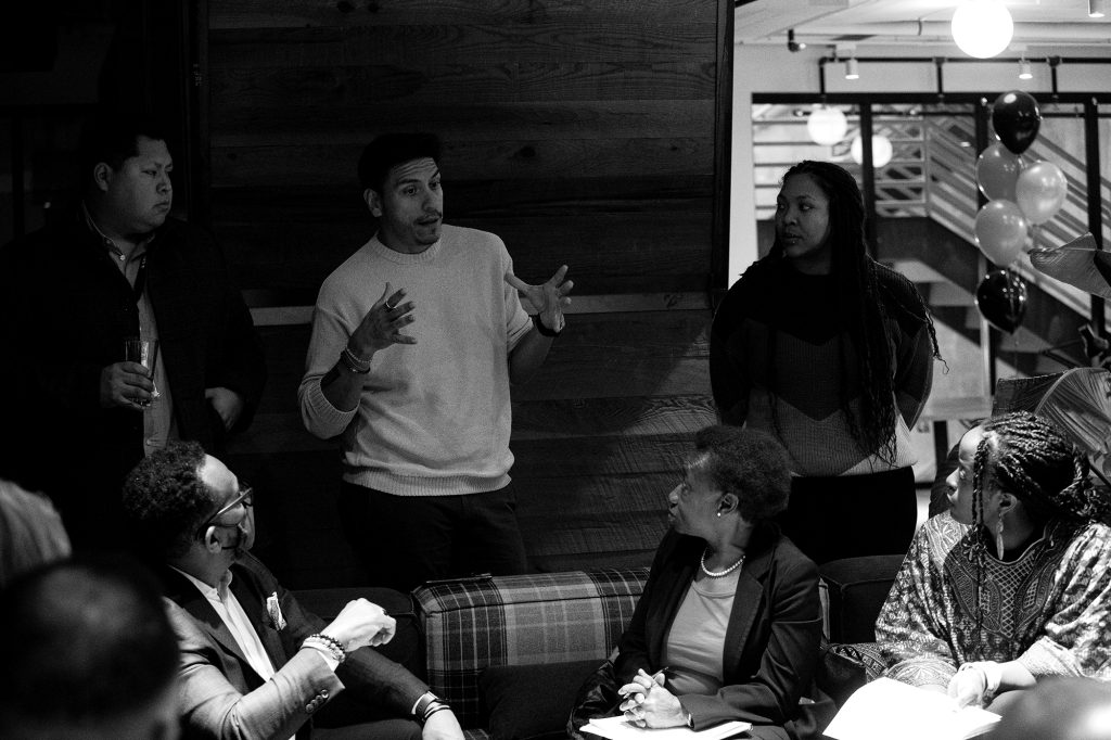 Balck and white image of Council Member Alejandro Puy speaking with seven people at the black history month annual community meeting.