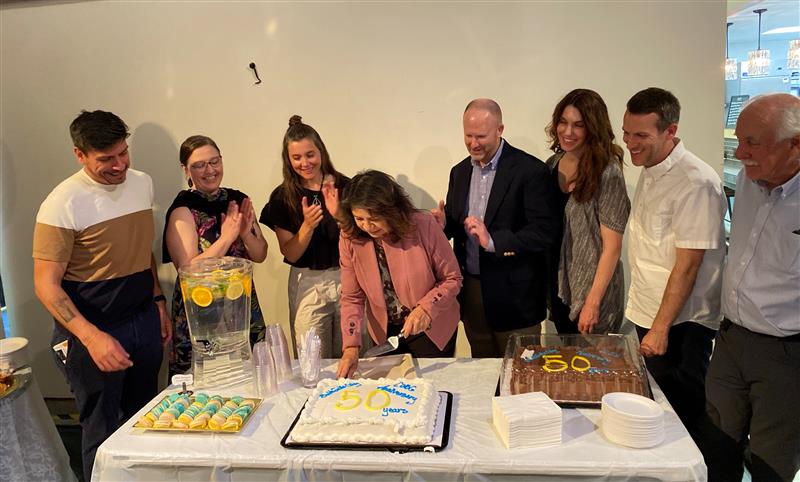 Councile Member Puy and six other peopls stand around a table while a woman in a pink jacket cuts a cake.