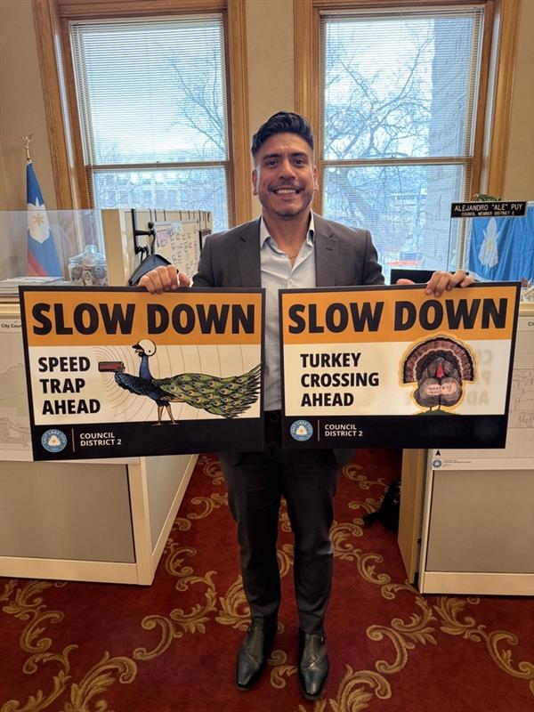 Council Member Ale Puy holding two “Slow Down” signs. One says “Slow Down Speed Trap Ahead” with a peacock dressed as a police officer holding a radar speed gun. The other says “Slow Down Turkey Crossing Ahead” with an illustration of a turkey with a tattoo saying "West $ide Pride." 