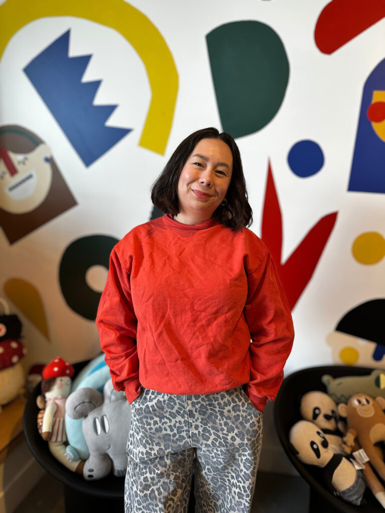 A woman named Amanda in a red top and gray leopard print pants stands in front of an abstract mural with colorful shapes.