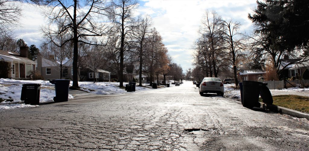 2500 East in winter, showing cracked pavement and surface wear, with snow lining the curbs and garbage bins placed for collection.