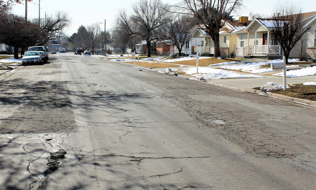 View of 1000 West before reconstruction, showing cracked and deteriorated pavement, snow patches on lawns, and parked cars lining the curb.