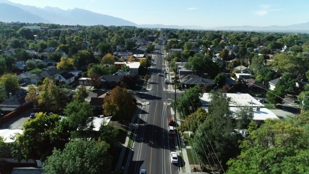 Aerial view of 1100 East in Salt Lake City lined with houses, trees and vehicles, with mountains in the background on a clear day.