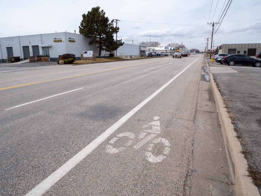 Existing conditions on 1700 South looking west at roughly 800 West. There are two travel lanes in each direction with a center two way left turn lane. There are also small painted bike lanes on the edges of the road in each direction. 