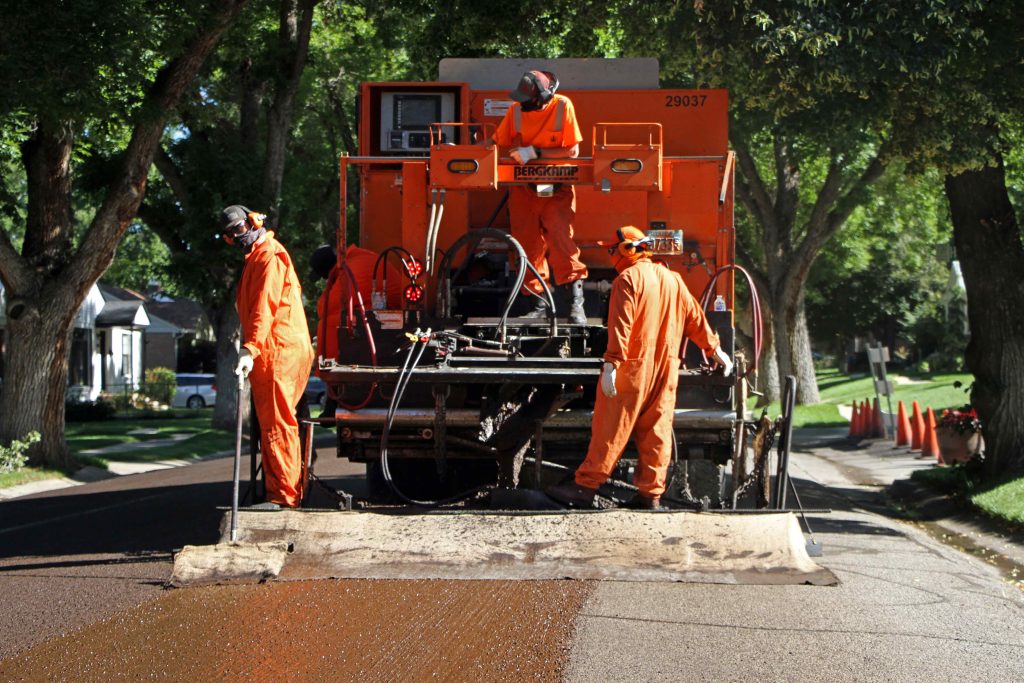 The SLC Streets team, dressed in orange jumpsuits and standing on a large asphalt truck, perform a slurry seal operation.