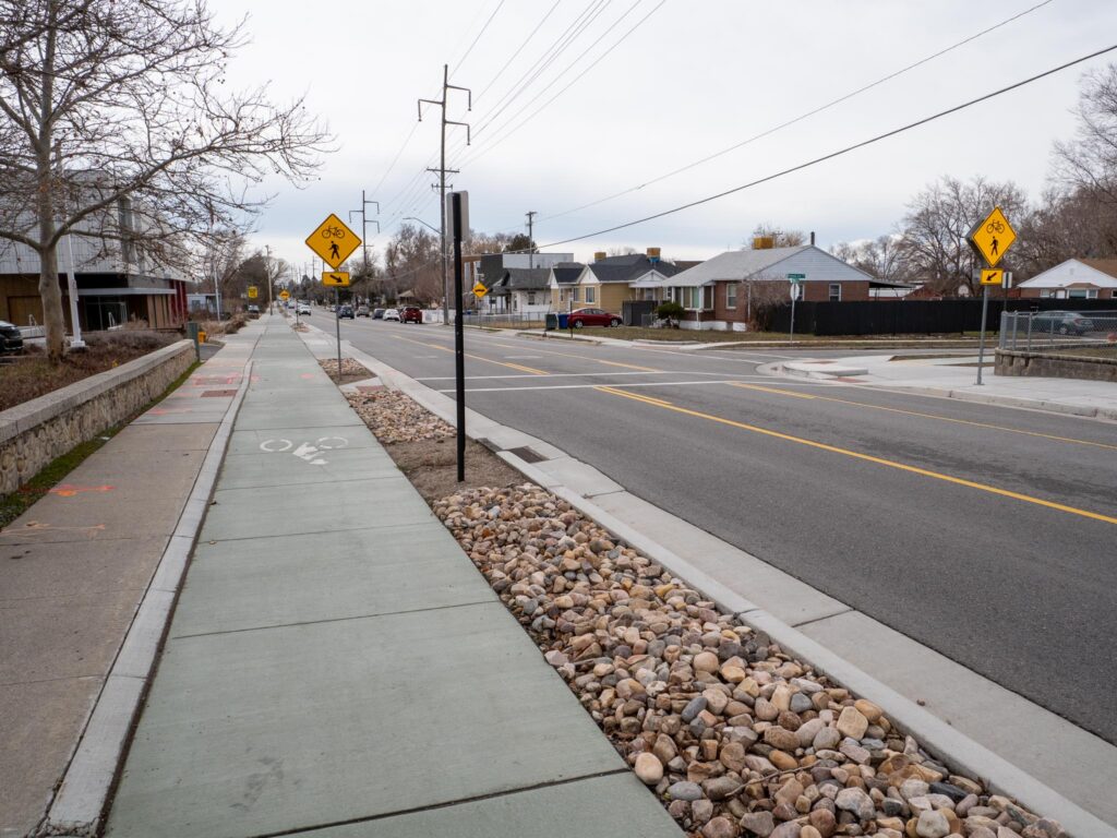 A street view of 900 East showing new walking and biking areas. A wide sidewalk and bike path sits next to the road, separated by a strip of rocks. There is also a marked crosswalk for crossing the street.