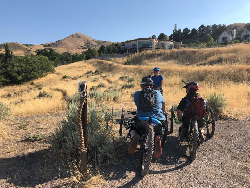two people on adaptive bikes wait on a trailhead