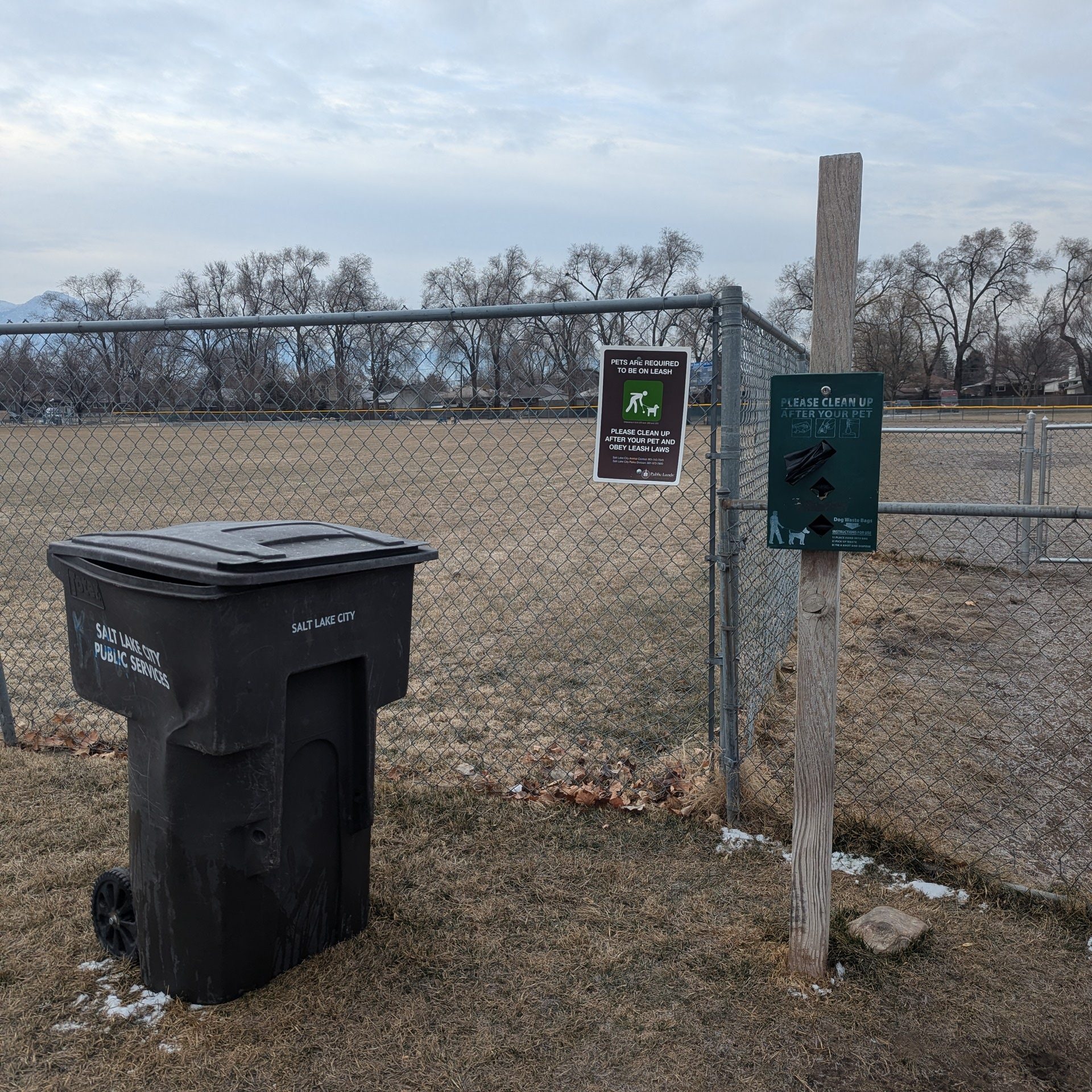 Photo shows one entrance to the off leash dog park, with dog waste bags, a trash can, and an air lock double gate