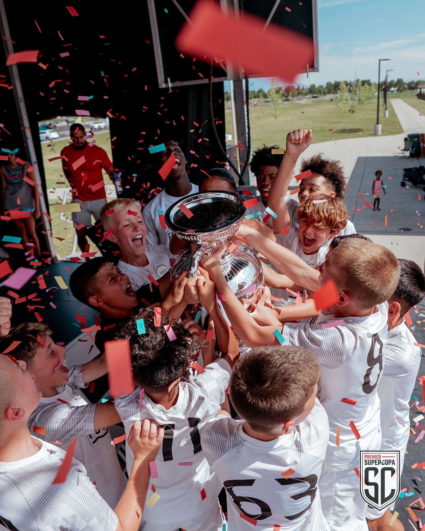 young boys in soccer uniforms cheer, holding a large trophy with confetti raining down