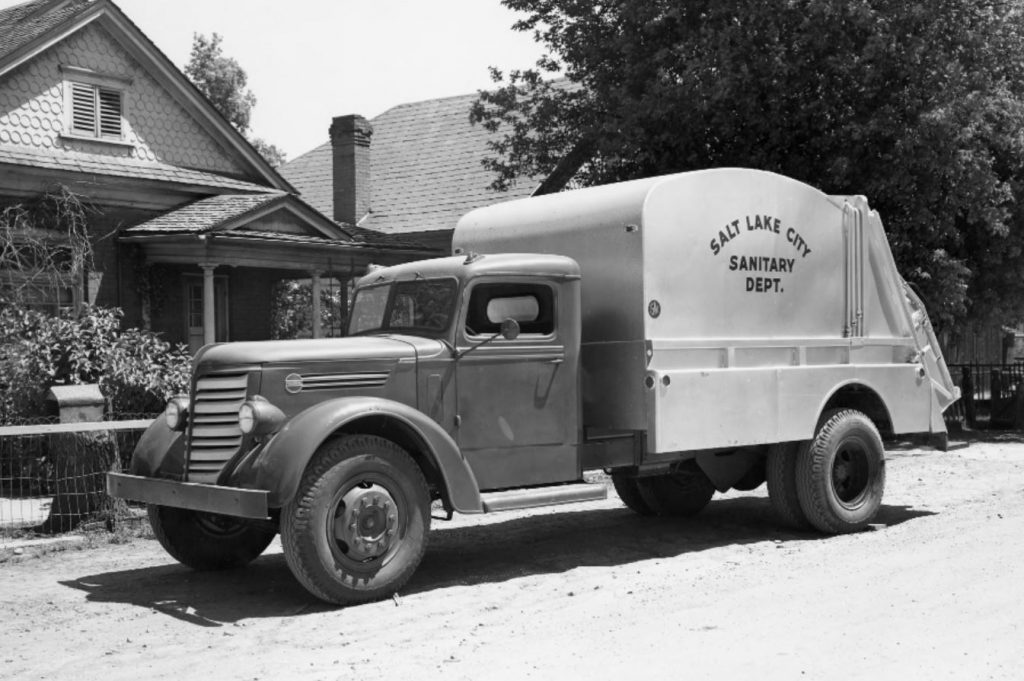 Photograph of Salt Lake City Sanitation Department garbage truck parked on unpaved street outside a small bungalow in Salt Lake City. 