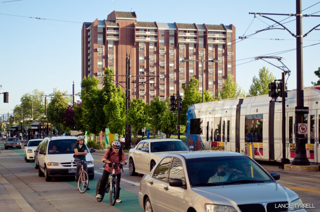 A picture of vehicles and cyclists waiting at a traffic signal on Main Street. TRAX light rail passes by in the background.