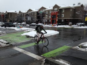 Cyclist on a red bike crossing a snowy urban intersection with green bike lane markings.
