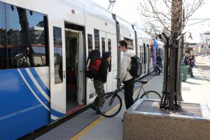 People at a TRAX light rail station with a train, bike, and a tree in a planter.