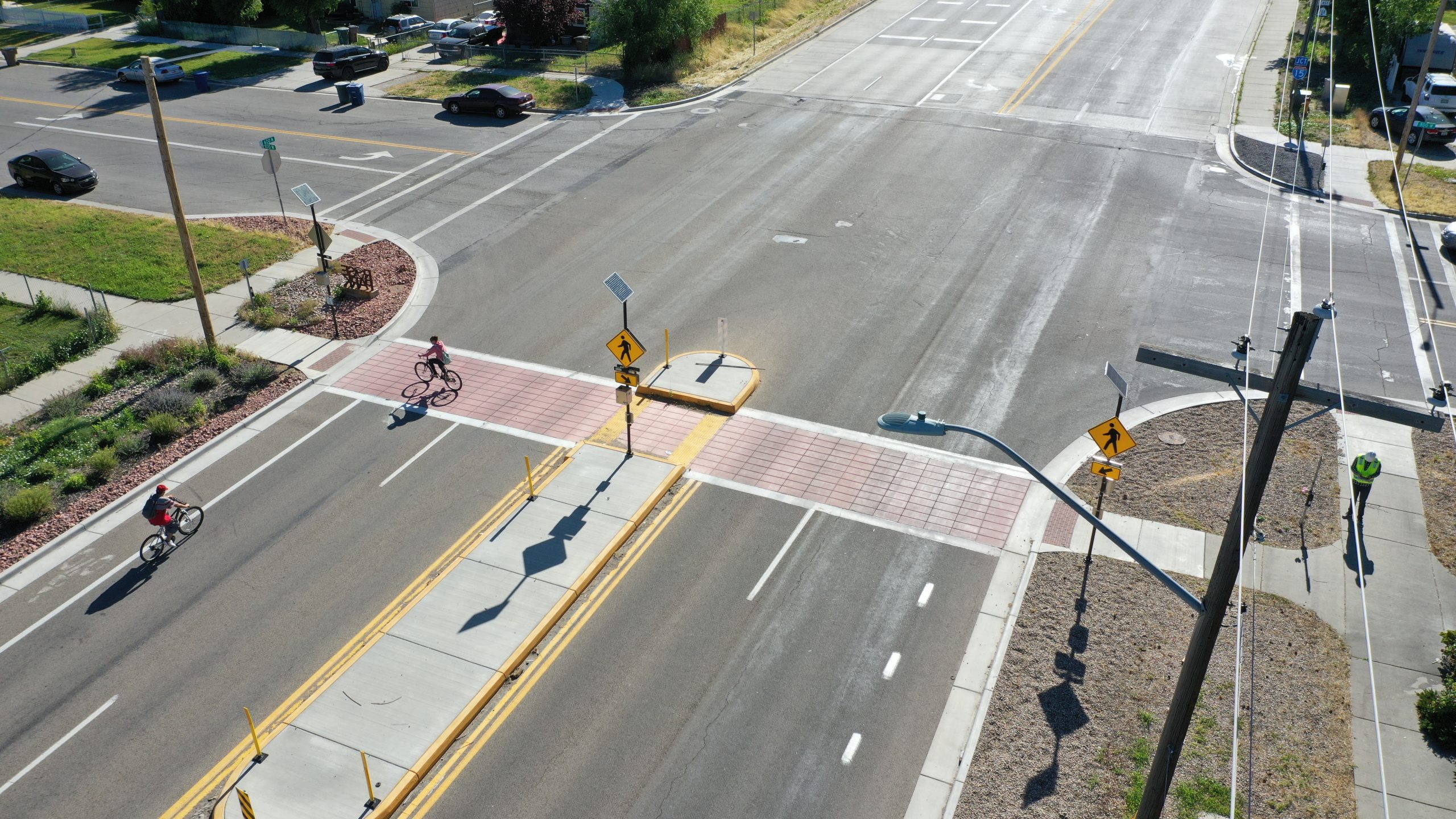 The 600 North 800 West intersection from a birds eye view, with a cyclist crossing the street. There is a red concrete crosswalk, a pedestrian refuge island, and flashing crosswalk signs on both sides of the street.