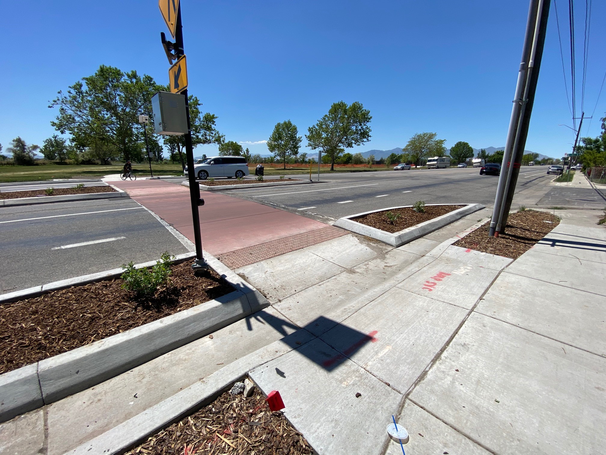A crosswalk improvement at 1300 West and 1700 South. The crosswalk was upgraded to red concrete to stand out against the black asphalt road. Pedestrian push buttons mounted to flashing yellow crosswalk signs are located on each side of the road. Concrete curb extensions and a pedestrian refuge island filled with mulch and shrubs shorten the crossing distance and make pedestrians more visible to drivers. 