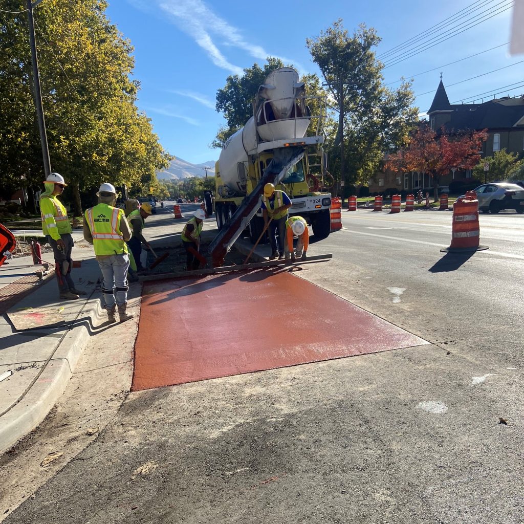 Construction workers pouring a red concrete pad within a black asphalt road.