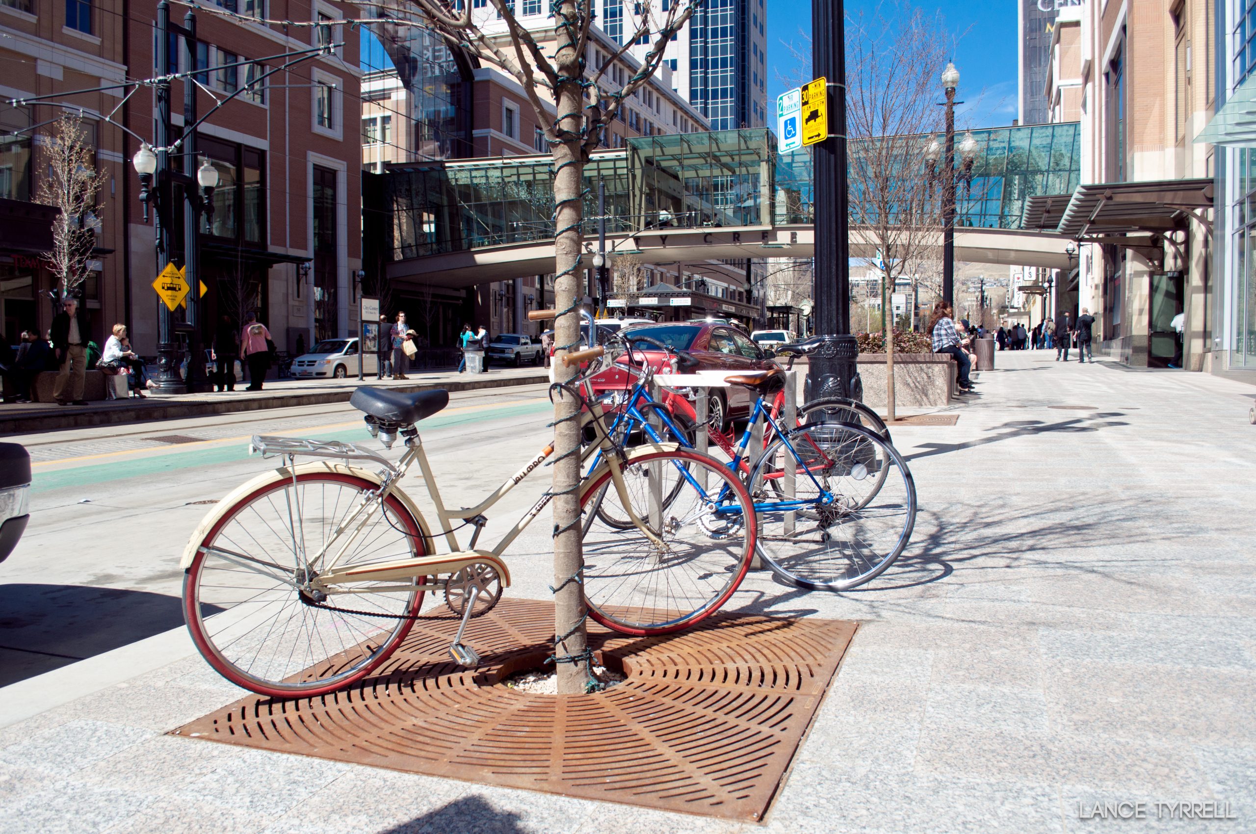 Two bicycles parked and secured to a tree on a city sidewalk near City Creek Center in downtown Salt Lake City. Pedestrians walk in the background, and buildings and a glass skybridge stretch above the street.