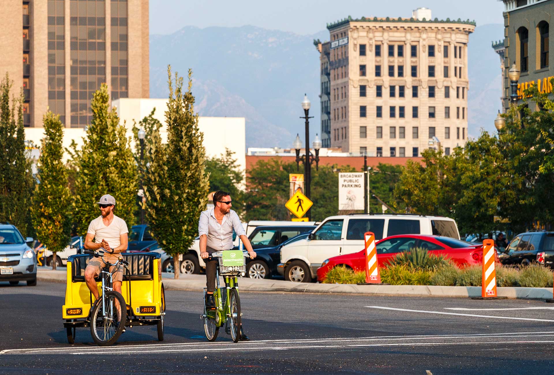 Two men are biking across a Salt Lake City street: one rides a GREENbike (bike-share) while the other pedals a yellow bike taxi. Tall buildings and trees line the background, with a mix of cars, pedestrians, and signage indicating a bustling urban setting.