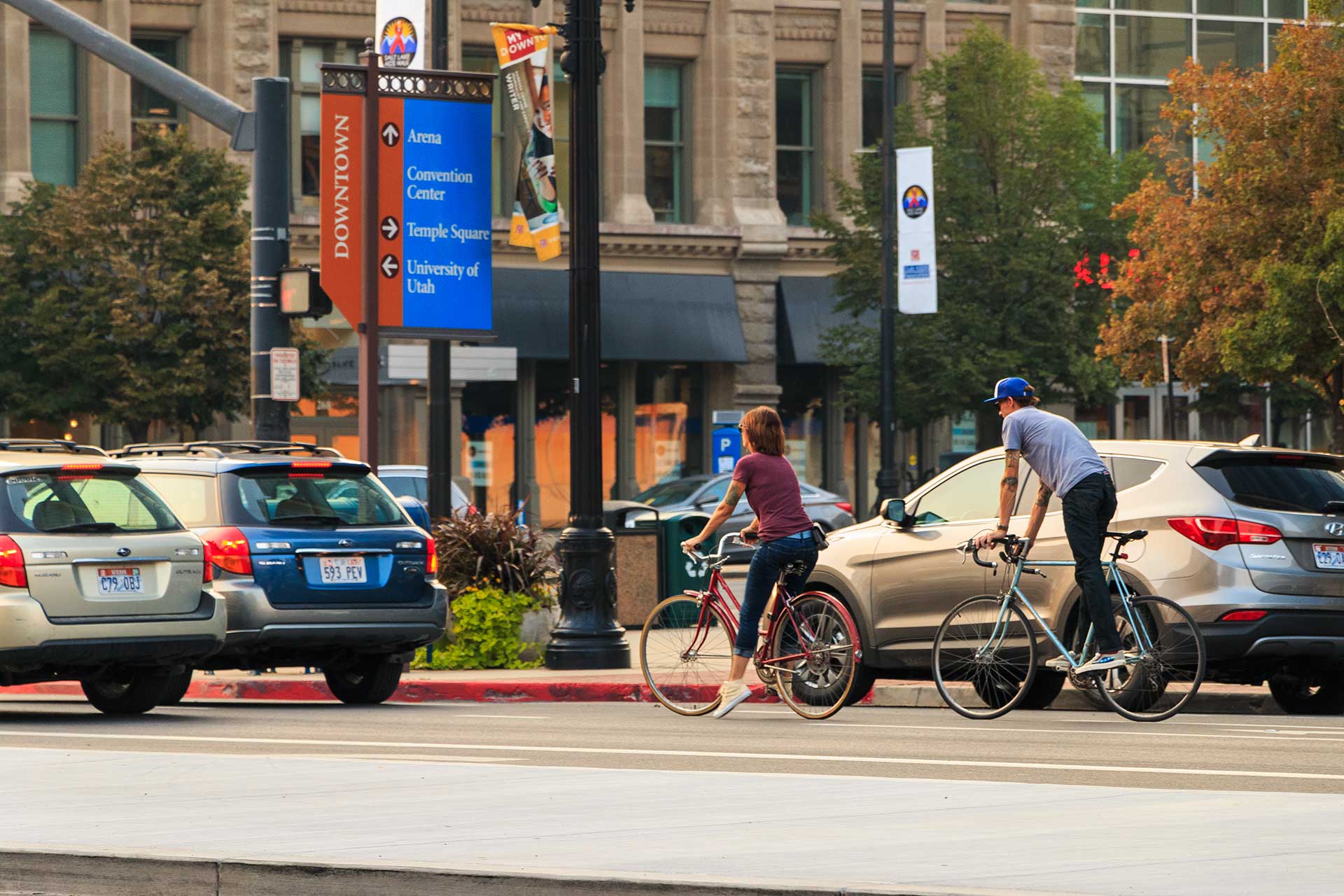 Two cyclists ride along a downtown Salt Lake City street. They pass a blue directional sign with arrows to nearby destinations like the Arena, Convention Center, and University of Utah. Behind them are parked cars, trees, and historic buildings, creating a lively urban atmosphere.