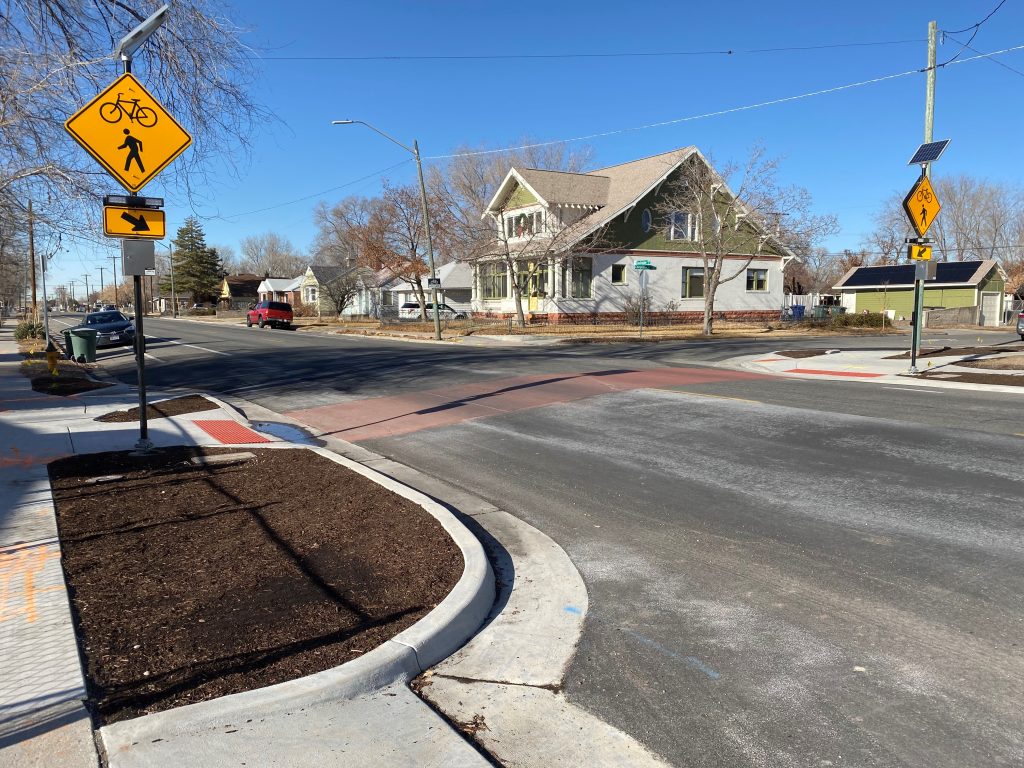 A crosswalk improvement at the intersection of Indiana Avenue and Cheyenne Street. The crosswalk was upgraded to red concrete to stand out against the black asphalt road. Pedestrian push buttons mounted to flashing yellow crosswalk signs are located on each side of the road. Concrete curb extensions filled with mulch shorten the crossing distance and make pedestrians more visible to drivers. The curb extensions have a separate ramp for bicycles to make it easier for bicycles to safely cross the street. 