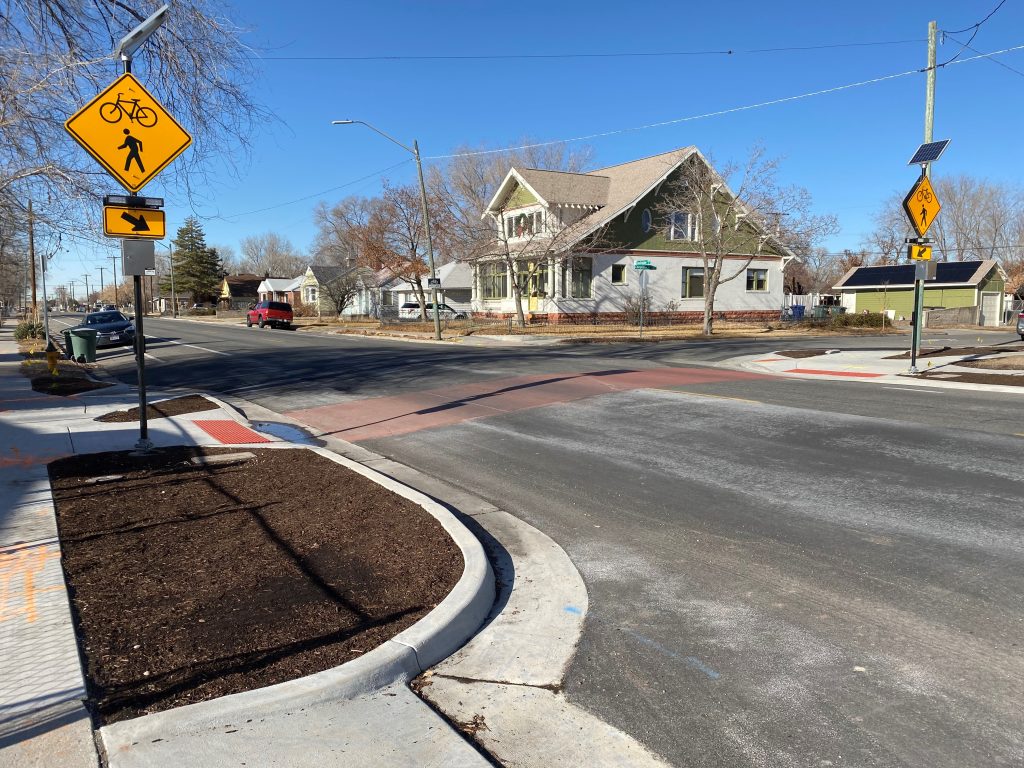 A crosswalk improvement at the intersection of Indiana Avenue and Cheyenne Street. The crosswalk was upgraded to red concrete to stand out against the black asphalt road. Pedestrian push buttons mounted to flashing yellow crosswalk signs are located on each side of the road. Concrete curb extensions filled with mulch shorten the crossing distance and make pedestrians more visible to drivers. The curb extensions have a separate ramp for bicycles to make it easier for bicycles to safely cross the street.