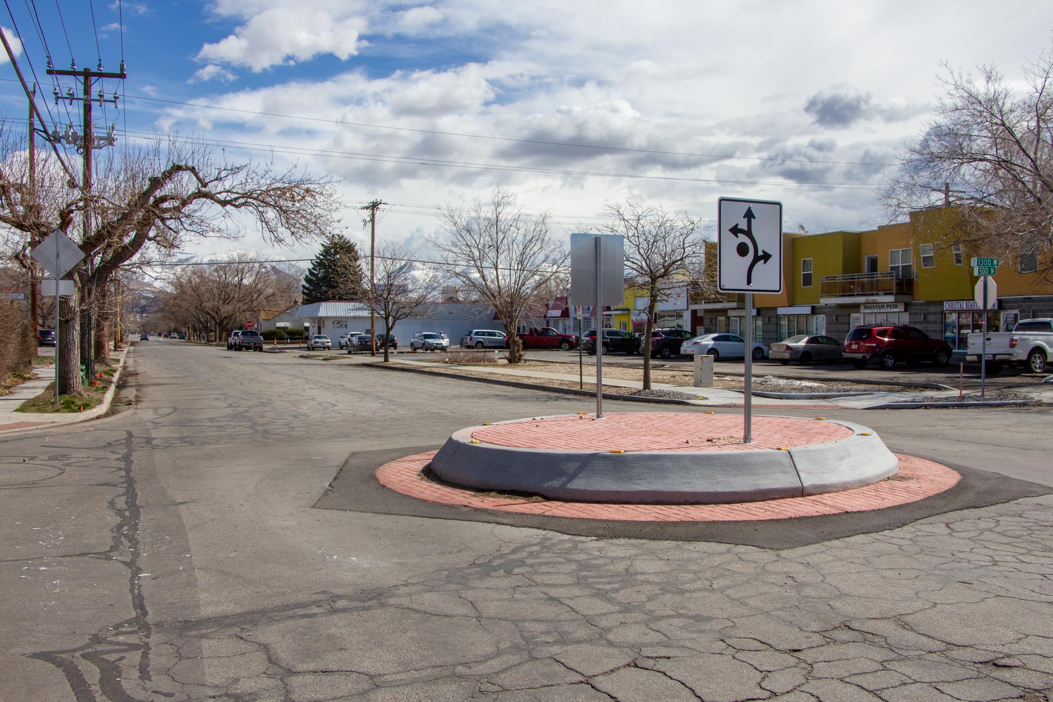 A traffic circle at the intersection of 1300 West and 500 North. It consists of a raised concrete ring with stamped red concrete on the flat top. Two signs that help with navigating the traffic circle are bolted into the concrete. 