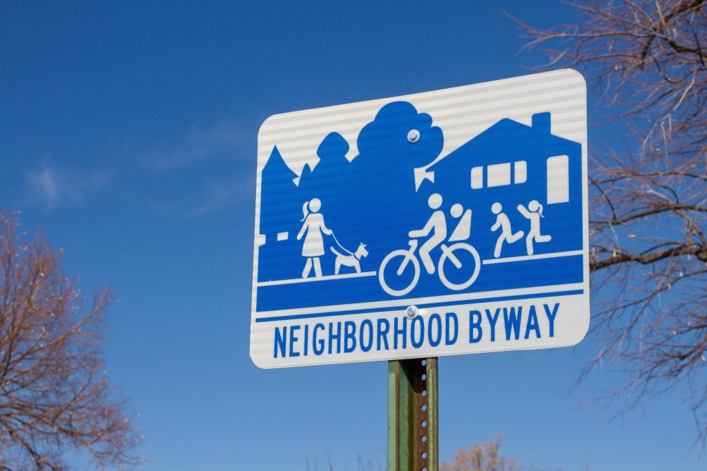 A blue and white Neighborhood Byway sign with people walking and biking in the street.