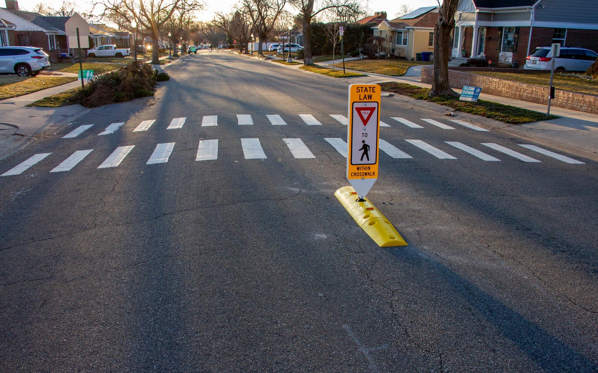 A small, yellow plastic curb adjacent a crosswalk in the street. Atop the curb, there is a tall, narrow sign that says "Yield to Pedestrians." The sign is mounted to a flexible delineator post.