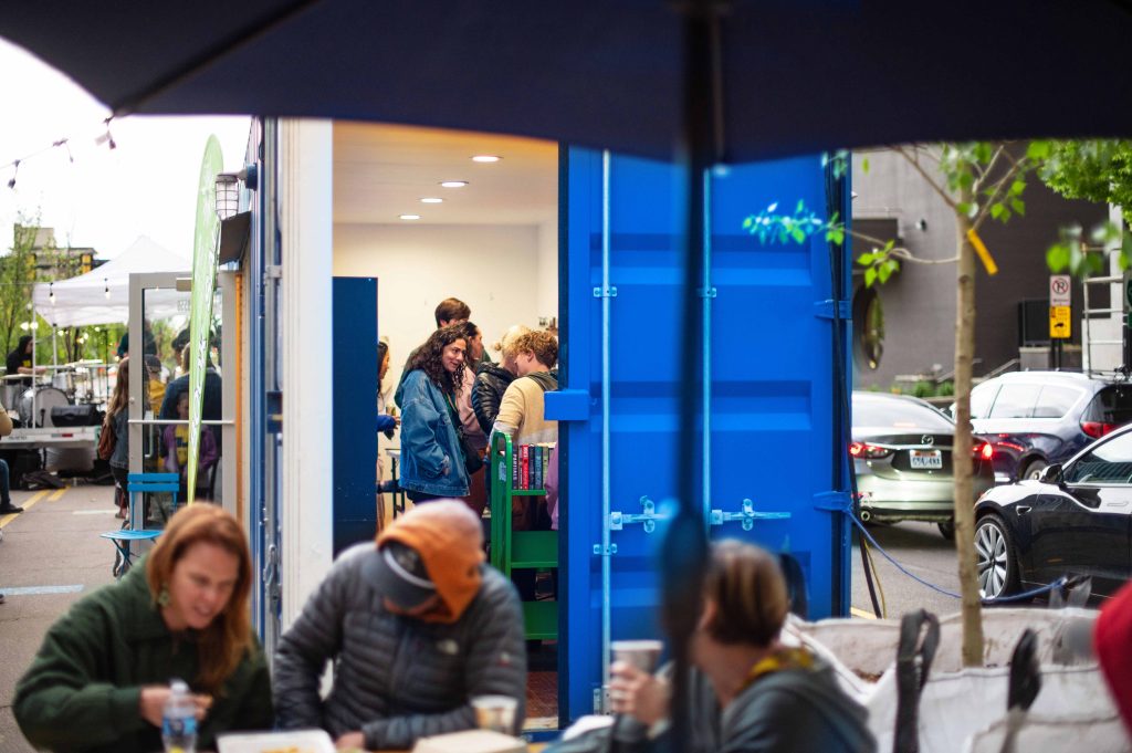 A group of people looking at books inside the book store at the Green Loop. The book store is inside of a bright blue shipping container.