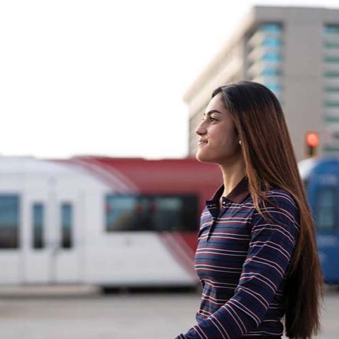 A woman with long brown hair in a striped shirt stands near a moving transit train in an urban area.